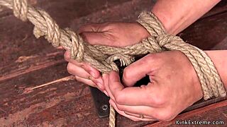 waxed participant endures back arch hogtie in session.