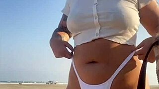 Redhead exhibitionist flashes swollen pussy lips on beach, muscular guy watches intently