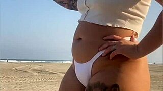 Redhead exhibitionist flashes swollen pussy lips on beach, muscular guy watches intently